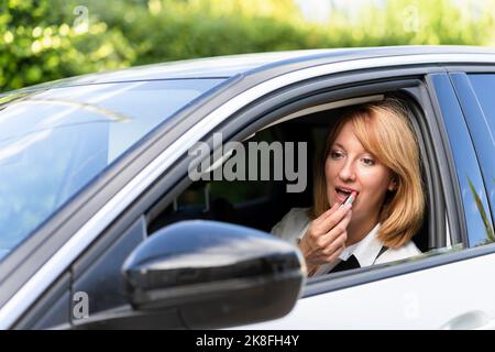 Mature woman looking at side-view mirror of car and applying lipstick Stock Photo