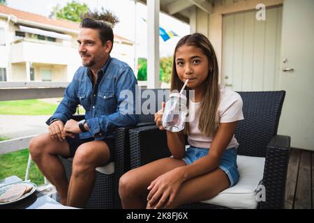 Daughter sipping drink sitting on chair with father at porch Stock Photo