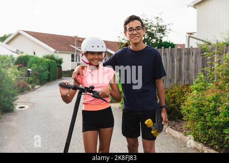 Smiling boy with arm around sister wearing helmet standing on road Stock Photo