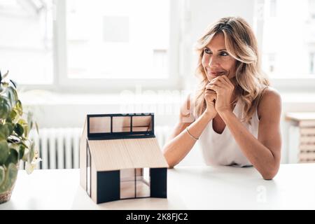 Smiling businesswoman day dreaming by house model at desk in office Stock Photo