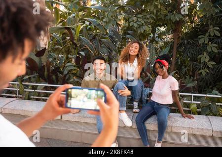 Young man photographing smiling friends through smart phone on wall Stock Photo