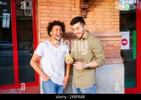 Young man laughing with eyes closed by friend using smart phone Stock Photo