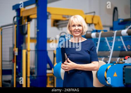 Happy senior businesswoman with tablet PC standing in front of machine Stock Photo