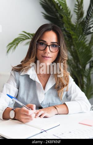 Smiling young beautiful lawyer with pen and book making notes at office Stock Photo