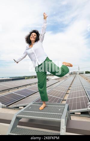 Happy businesswoman with hand raised in front of solar panels on rooftop Stock Photo