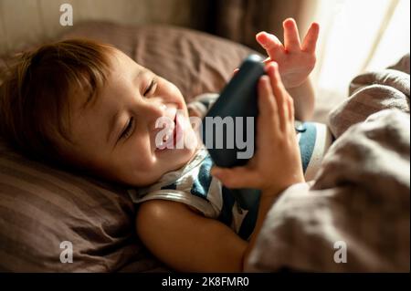 Smiling toddler using mobile phone lying with blanket in bed at home Stock Photo