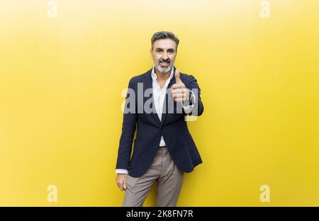 Smiling businessman showing thumbs up gesture against yellow wall Stock Photo