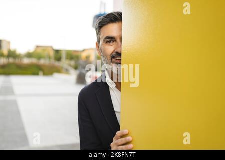 Smiling businessman peeking behind yellow wall Stock Photo