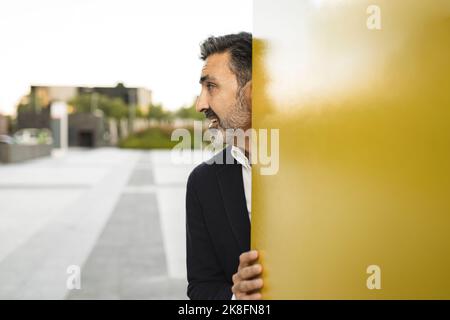 Playful businessman hiding behind yellow wall Stock Photo