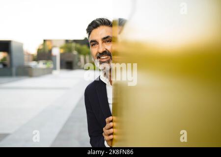 Happy businessman hiding behind yellow wall Stock Photo
