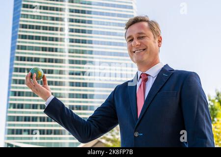 Smiling businessman holding small globe ball in front of office building Stock Photo