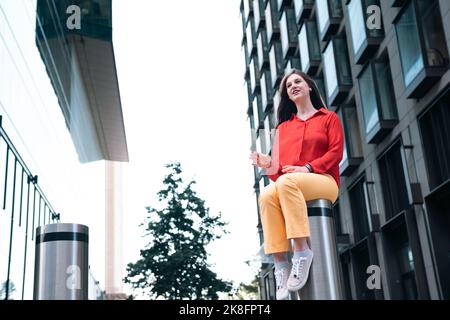 Young woman sitting on bollard in front of building Stock Photo