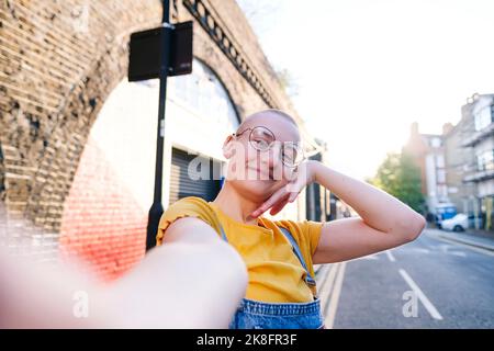 Smiling non-binary person with shaved head taking selfie on street Stock Photo