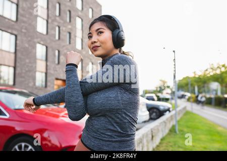 Young woman listening to music and doing stretching exercise on footpath Stock Photo