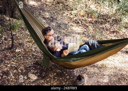 Grandfather reading book for grandson lying on hammock in forest Stock Photo