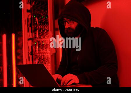 Hacker wearing hoodie doing coding on laptop in server room Stock Photo