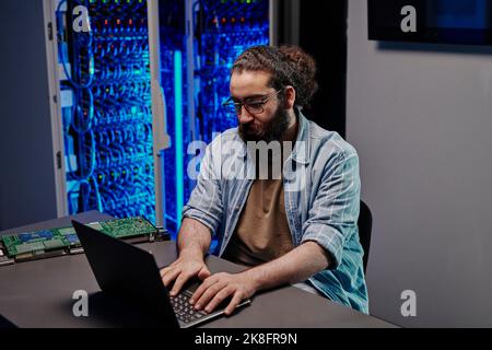 IT expert working on laptop sitting at desk in server room Stock Photo