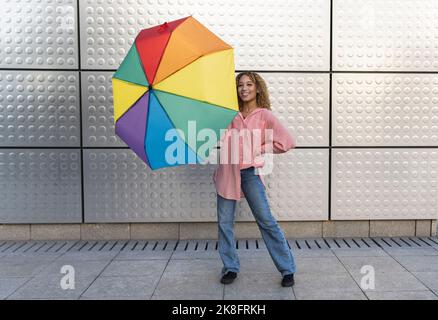 Smiling young woman with rainbow umbrella on footpath Stock Photo