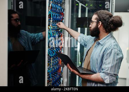 Technician connecting cable on server holding laptop in server room Stock Photo