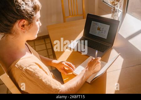 Freelancer taking down notes in diary by laptop at desk Stock Photo