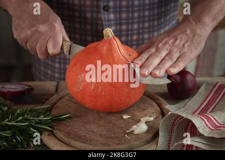 Crop anonymous male cutting fresh pumpkin with sharp knife on wooden cutting board in kitchen Stock Photo