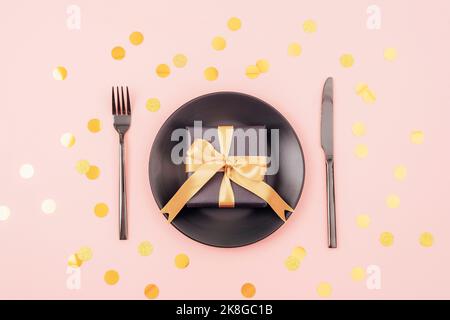 Stylish table setting, gift box, black plate, cutlery on pink background with golden confetti. Christmas concept. Top view, flat lay Stock Photo