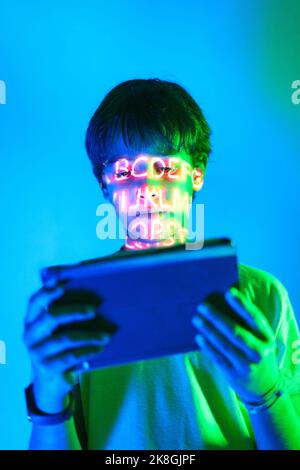 Preteen kid with short hair standing in room with neon illumination inscription and playing online videogame via tablet Stock Photo