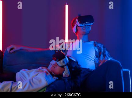 Multiracial unrecognizable couple in VR goggles sitting together on couch and interacting with virtual reality in dark room Stock Photo