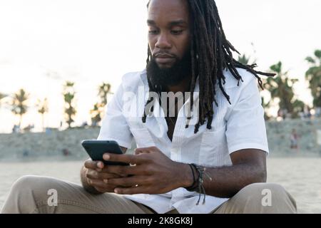 Concentrated African American male with dreadlocks browsing mobile phone while sitting on sandy beach against blurred background of trees Stock Photo