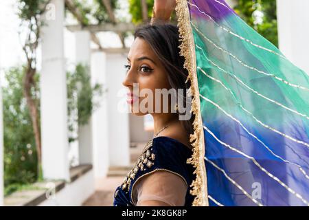 Side view of young Indian female in traditional clothes and headscarf smiling and dancing and looking away in green park Stock Photo