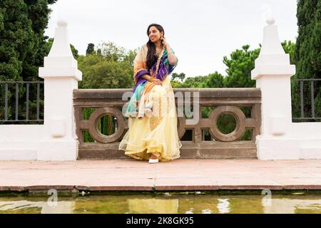 Full body of young Indian female in elegant dress standing near pond with green trees and bushes in background Stock Photo