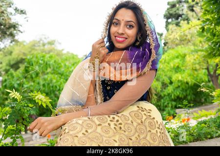 Young cheerful ethnic Indian female in stylish traditional sari and jewelry standing looking at camera in green park Stock Photo