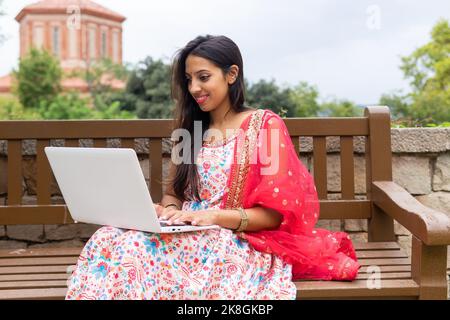 Young Indian female freelancer in dress sitting on wooden bench and typing on laptop while working on remote project Stock Photo