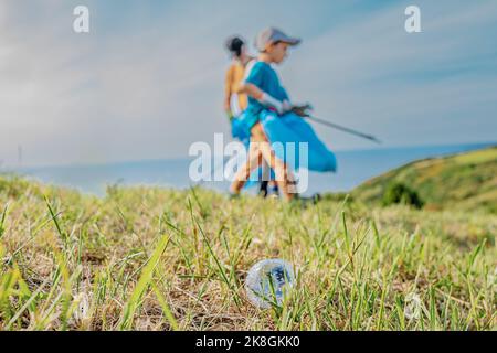 Empty used plastic bottle on grass with unrecognizable volunteers collecting garbage in background during environmental campaign in countryside Stock Photo