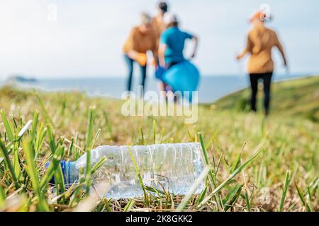 Empty used plastic bottle on grass with unrecognizable volunteers collecting garbage in background during environmental campaign in countryside Stock Photo