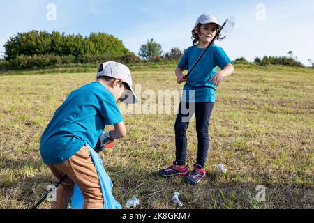 Preteen volunteers in blue t shirts and caps with thrash tongs and bin bag while collecting plastic waste on grassy lawn Stock Photo