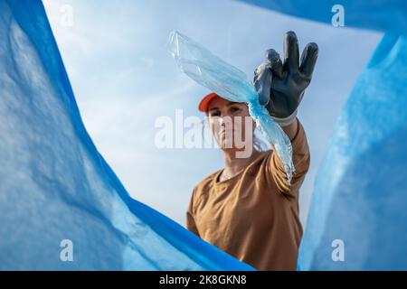 Low angle of smiling female volunteer in cap and protective gloves throwing garbage into blue bin bag while looking at camera Stock Photo