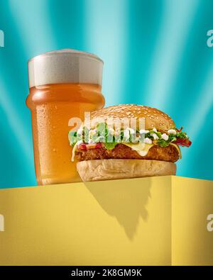 Delicious burger with fresh bun placed on yellow block near glass of cold foamy beer against turquoise background Stock Photo