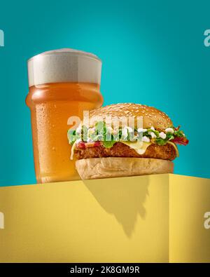 Delicious burger with fresh bun placed on yellow block near glass of cold foamy beer against turquoise background Stock Photo