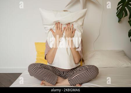 Unrecognizable woman in pajamas sitting on comfortable bed with crossed legs and hugging soft pillow while hiding from camera Stock Photo