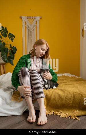 Positive ginger woman in pajamas sitting with eyes closed on comfortable bed tenderly caressing adorable cat with white paws Stock Photo