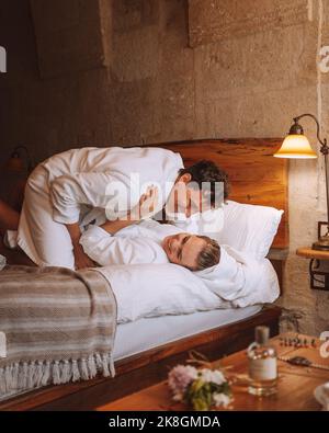 Cheerful young enamored couple in white bathrobes smiling happily while lying on bed and embracing each other in cozy hotel room Stock Photo