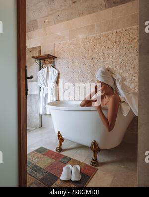 Side view of young female smiling with white towel on head while taking bath in light bathroom Stock Photo