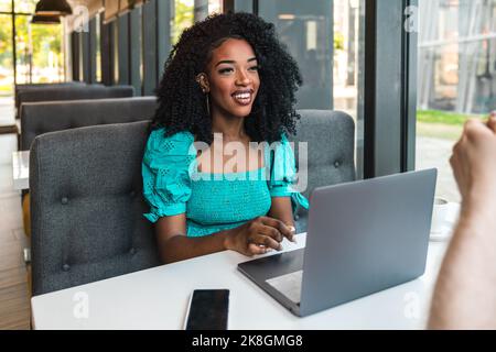Cheerful young African American woman in turquoise top sitting at table with smartphone and working via netbook while chatting with cropped unrecogniz Stock Photo