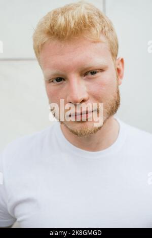 Pensive bearded adult blond haired man in white t shirt standing near white tiled wall looking at camera Stock Photo