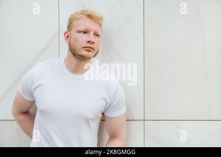 Pensive bearded adult blond haired man in white t shirt standing near white tiled wall looking away Stock Photo