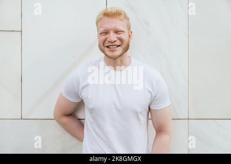 Cheerful bearded adult blond haired man in white t shirt standing near white tiled wall looking at camera Stock Photo