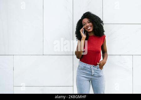 Smiling young ethnic woman in casual outfit standing near white tiled wall and holding hand in pocket while speaking on cellphone and looking away Stock Photo