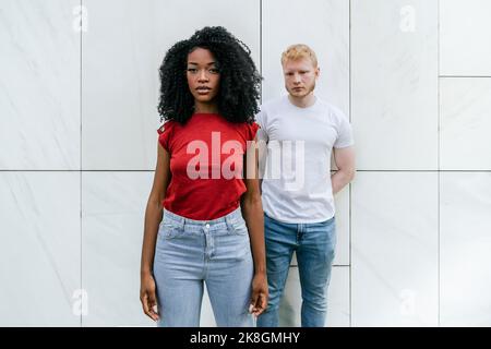 Pensive bearded blond man leaning on white tiled wall of building with hands behind back while standing next to African American woman in red t shirt Stock Photo