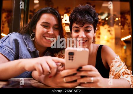 Happy young girlfriend smiling and browsing social media on cellphone while sitting at table during date in street cafeteria in Madrid, Spain Stock Photo